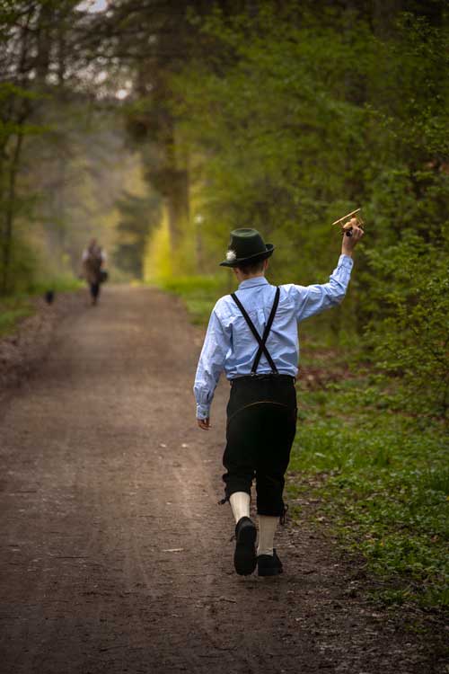 Portrait of a young boy holding an airplane in the air as it he were flying it himself. He wears traditional Bavarian clothing, lederhosen, trachten shoes and a hat.